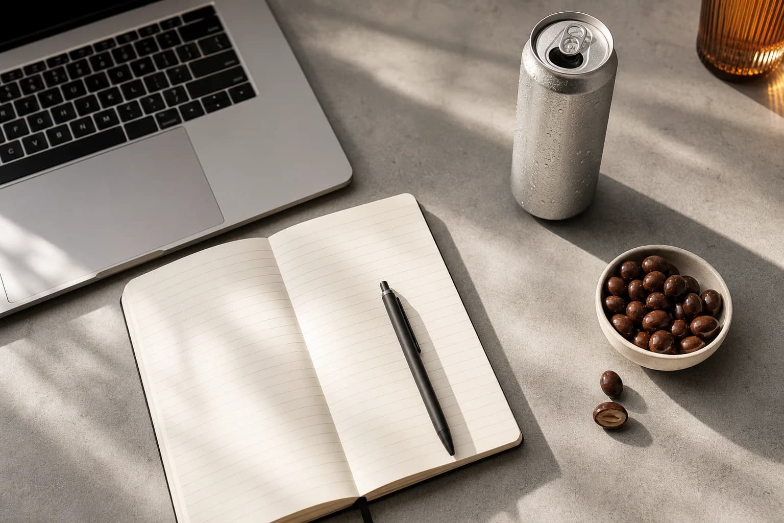 A desk flat lay with a notebook, laptop, energy drink can and a bowl of chocolate covered nuts